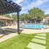 Green grass, brick and cement picnic table under a gazebo in the forground.  Pool with lounge chairs, trees in background.