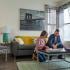 Block75 Apartments, interior, living room, man and woman playing board game, gray couch, chair, coffee table, wall art, wood floor, area rug, large windows, natural light