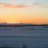 A view of an open, snowy field stretches out into a forest. The sun is setting above mountains in the distance. | Fort Greely, DOD housing