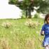 A young child in a blue dress standing in a field blowing on a dandelion.