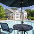 View of pool below umbrella with table and two chairs. Lounge chairs surround pool and clubhouse can be seen in background surrounded by trees