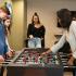 Block75 Apartments, interior, game room, one man and three women playing at foosball table, fourth woman watching and smiling, wall mounted tvs, seating area
