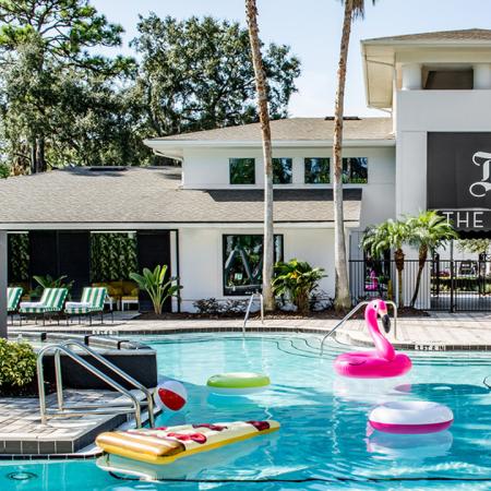 1800 The Ivy, exterior, sparkling blue swimming pool, club house entrance, lounge chairs, pink blow up flamingo, water toys, sign 'The Ivy' on side of gray and white building, trees,