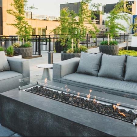 Fire pit lit up in front of 2 grey couches surrounded by greenery installations on the rooftop at Modera Broadway apartments.