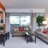 Clarke Manor Apts, interior, Living room, plush carpet, orange accent wall, large tan sofa and tan loveseat, coffee table, sliding glass door to patio, wall cut out into kitchen, central A/C vent,