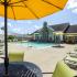View from table with yellow umbrella overhead towards pool with clubhouse located behind pool. Green lounge chairs surround pool on deck with umbrella covered tables and chairs. White privacy fence surrounds pool area. at Summit Place apartments in Methuen, MA.